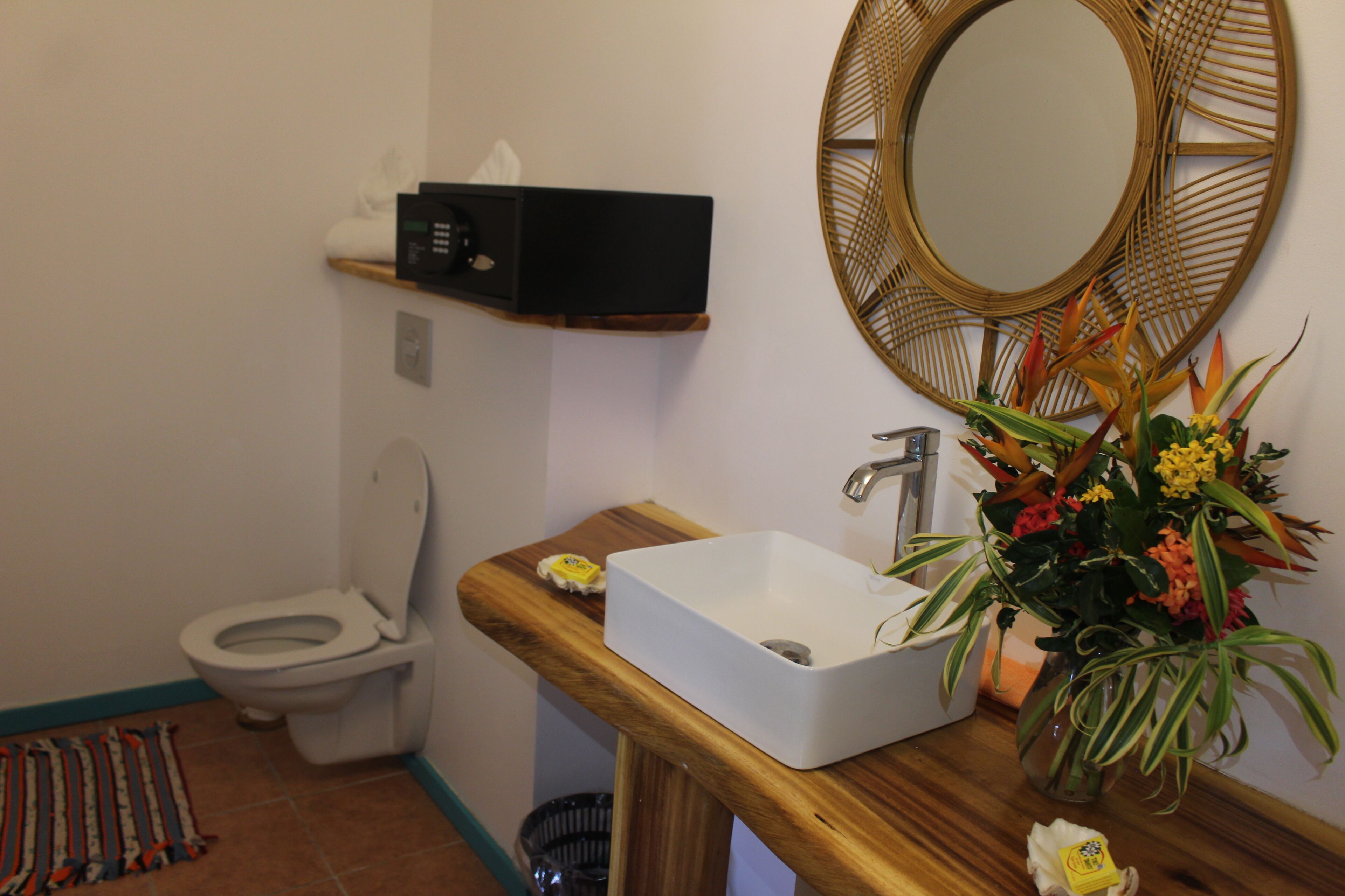 Un baño con un inodoro blanco, un tocador de madera con un lavabo blanco, un espejo y un florero con flores.
