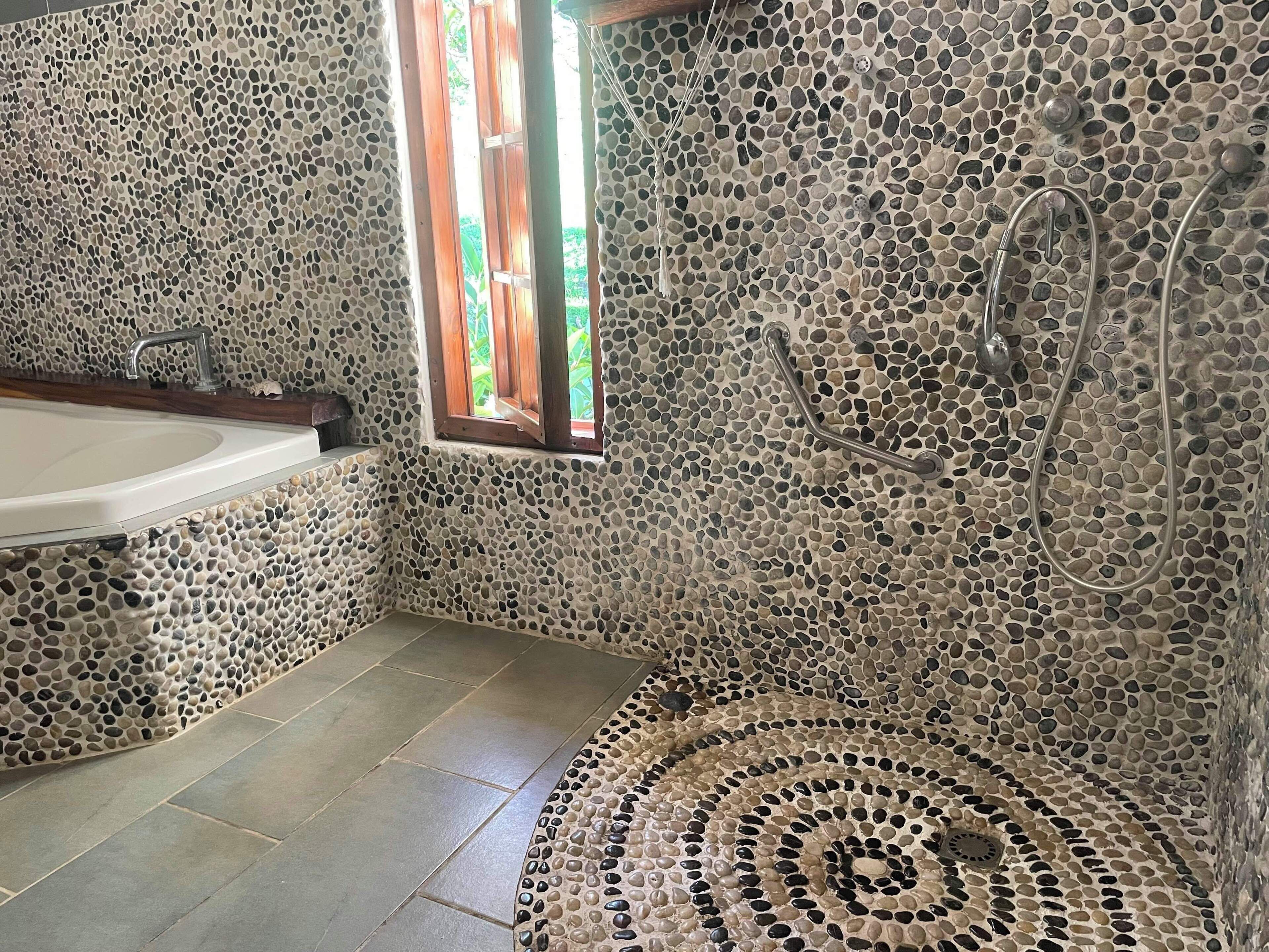 Ein Badezimmer mit Wand aus Steinmosaik, einer Badewanne, einer Dusche mit Duschbrause und einem Fenster mit Holzrollläden.