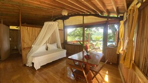 A wooden room with a bed, a table with a vase, and a view of greenery outside. - Fare Oviri Lodge (Raiatea)