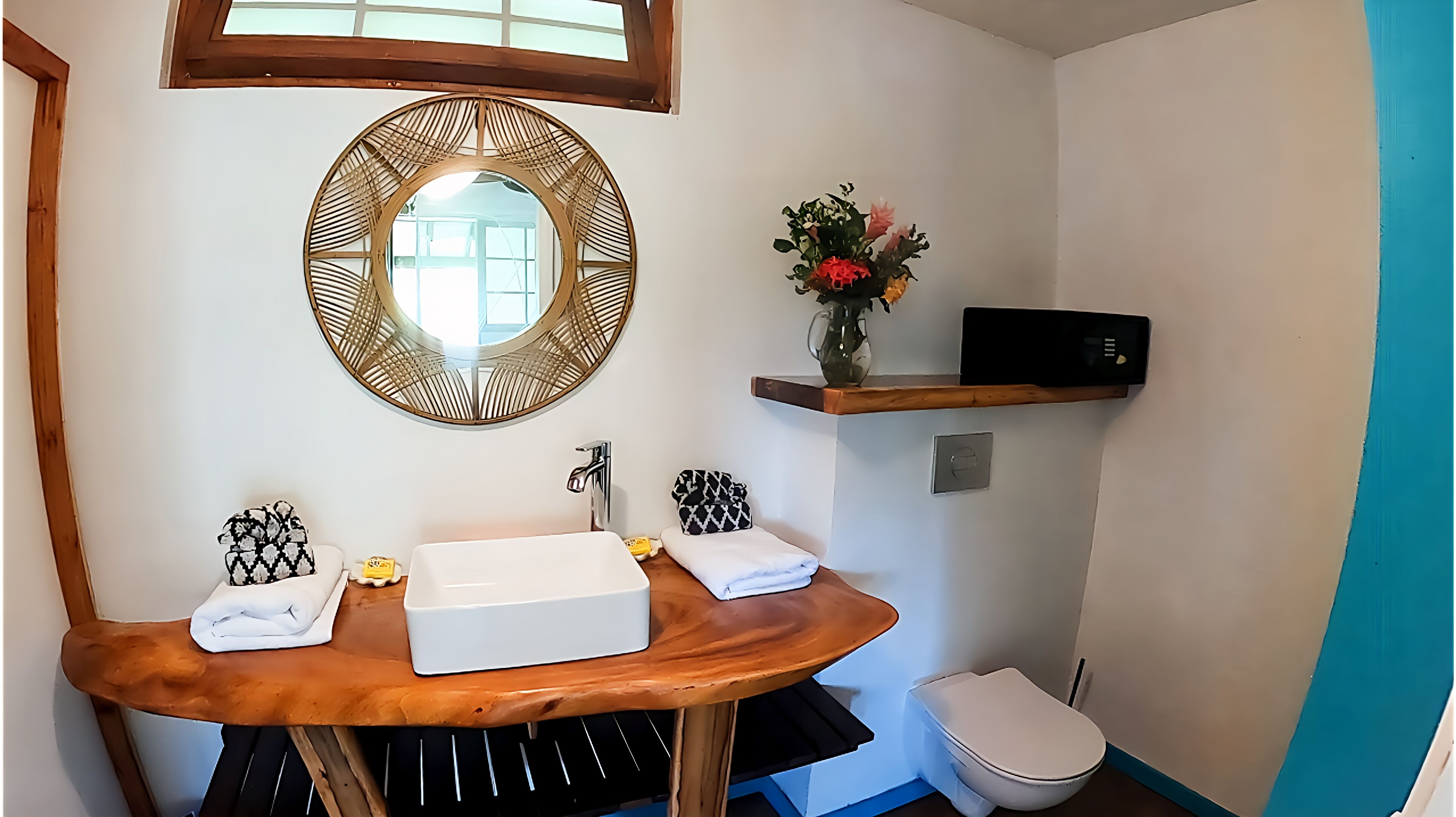 A bathroom with a wooden vanity, a round mirror, a shelf with a vase of flowers, a toilet, and a black microwave.