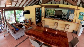 A spacious living area with a large wooden dining table, comfortable seating, and a kitchen area visible through an open doorway.