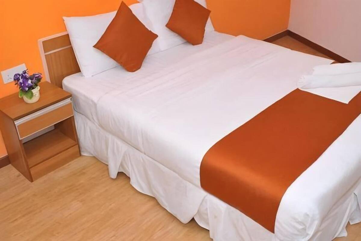 Una cama bien tendida con ropa de cama blanca y naranja, una mesita de noche de madera con un jarrón de flores y un suelo de madera.