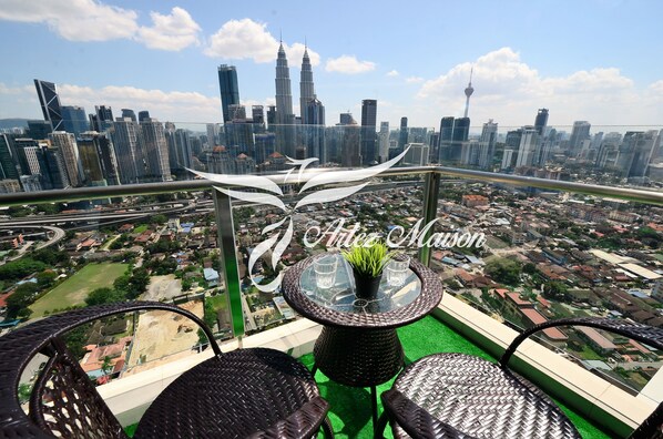 A balcony with wicker furniture, a glass table with a plant, and a cityscape view including the Petronas Towers.