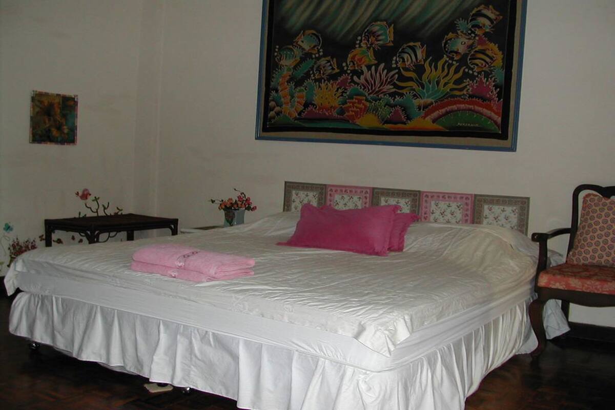 Una cama bien hecha con una colcha blanca y almohadas rosas, un cabecero de madera, una mesita con un florero y una silla con un cojín floral.