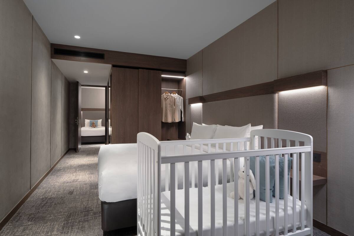 A modern hotel room with a crib, a bed, a walk-in closet, and a bathroom.