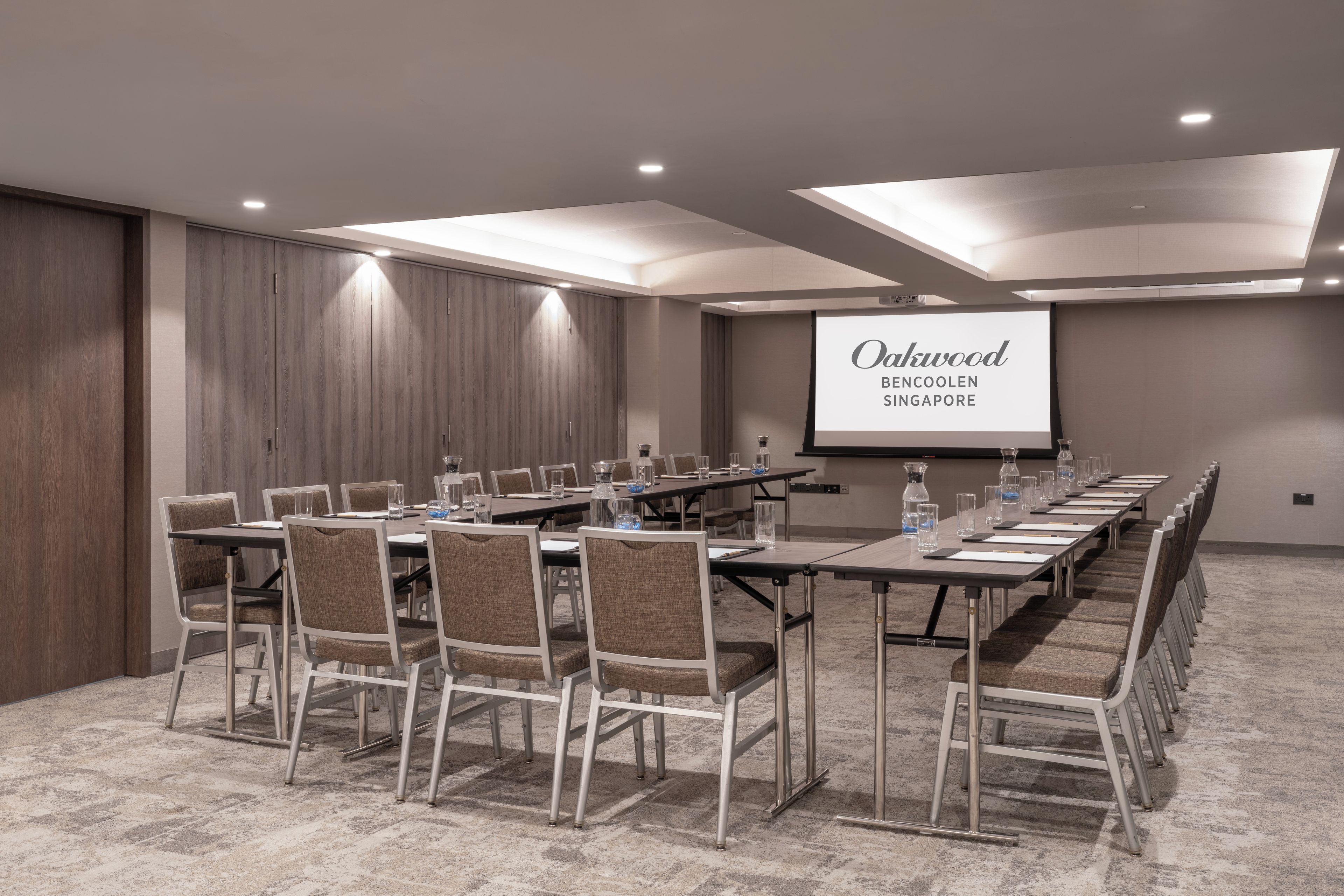 Un salón de conferencias con una larga mesa, sillas, una pantalla de proyector y suelo alfombrado.