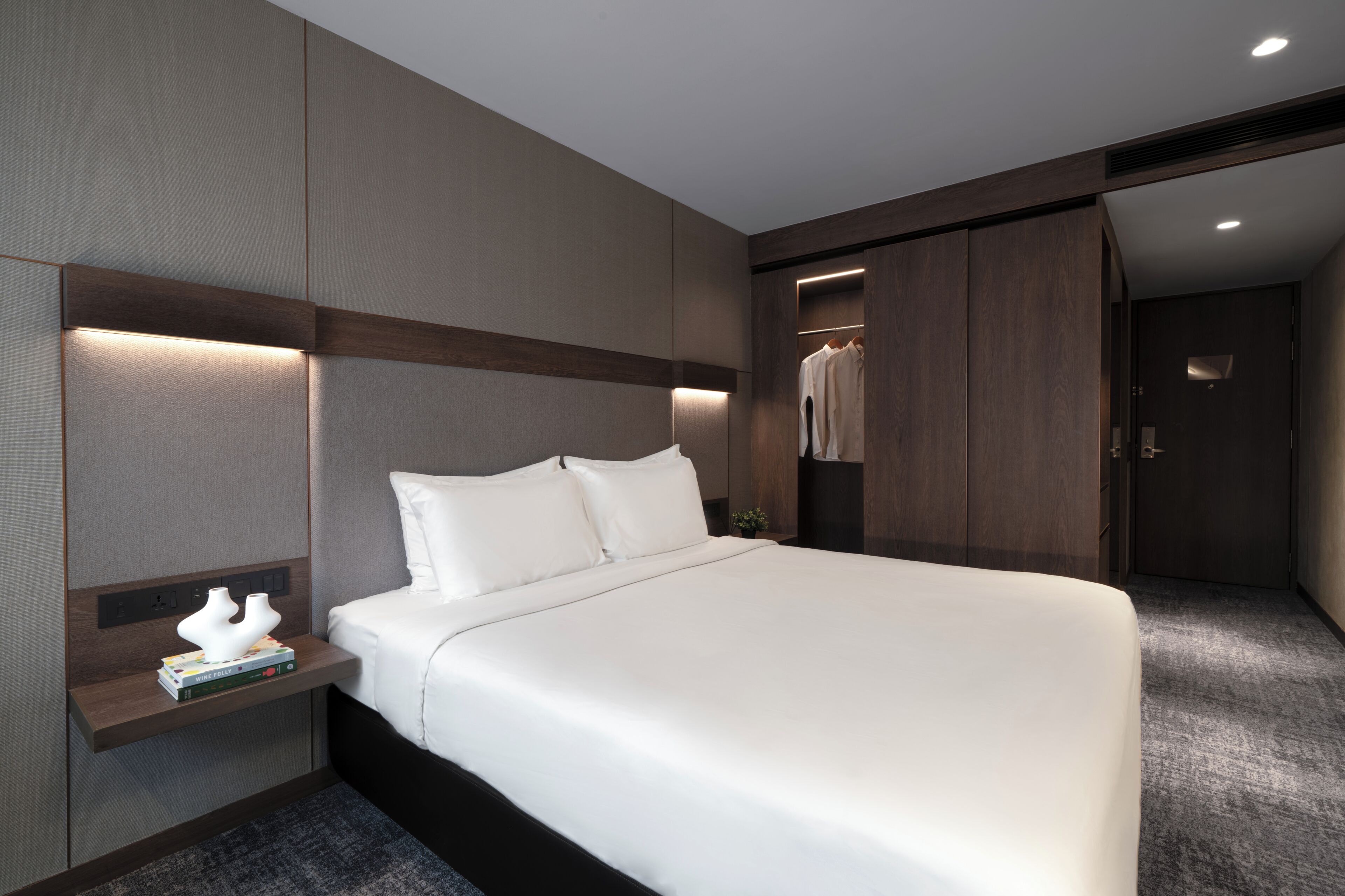 A modern hotel room with a large bed, a bedside table with books, a wardrobe, and a mirror.