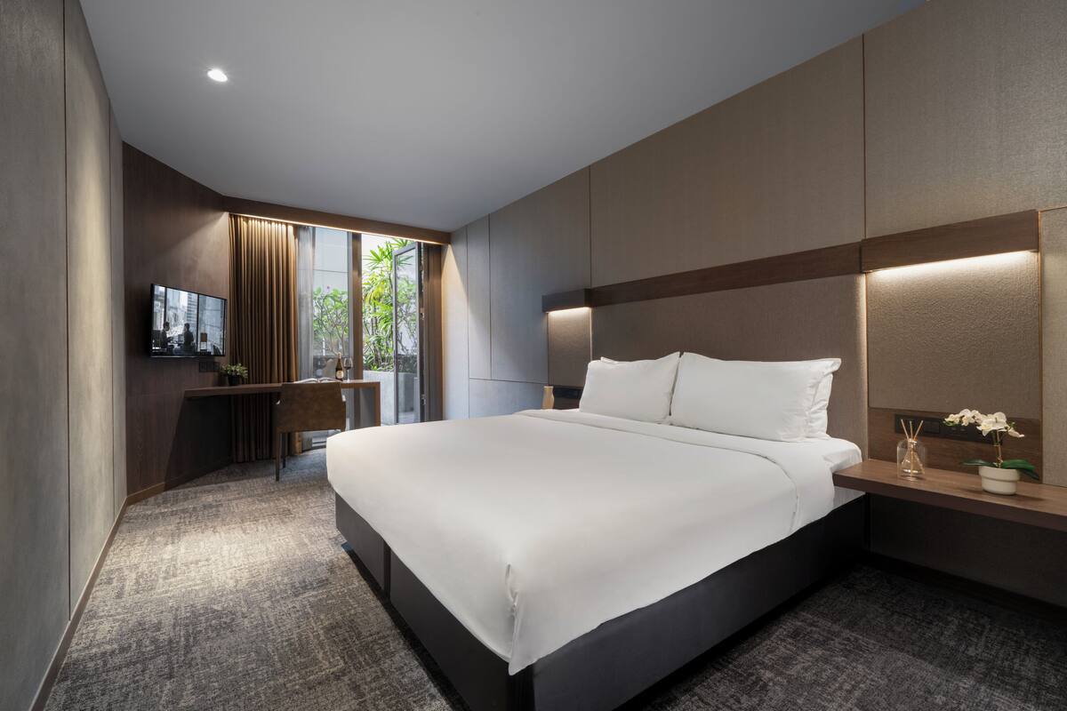 A modern hotel room with a large bed, a desk with a flat-screen TV, and a view of greenery outside.