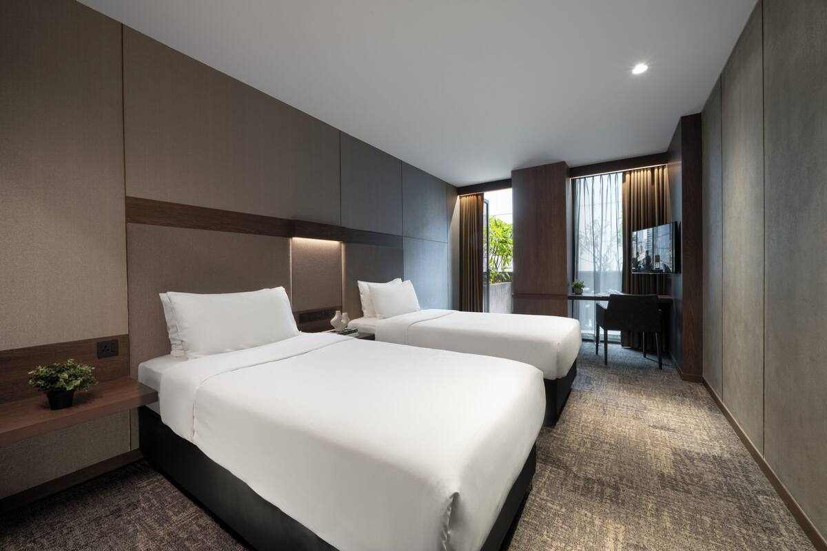 A hotel room with two beds, a desk, a chair, a small plant, and a window with curtains.