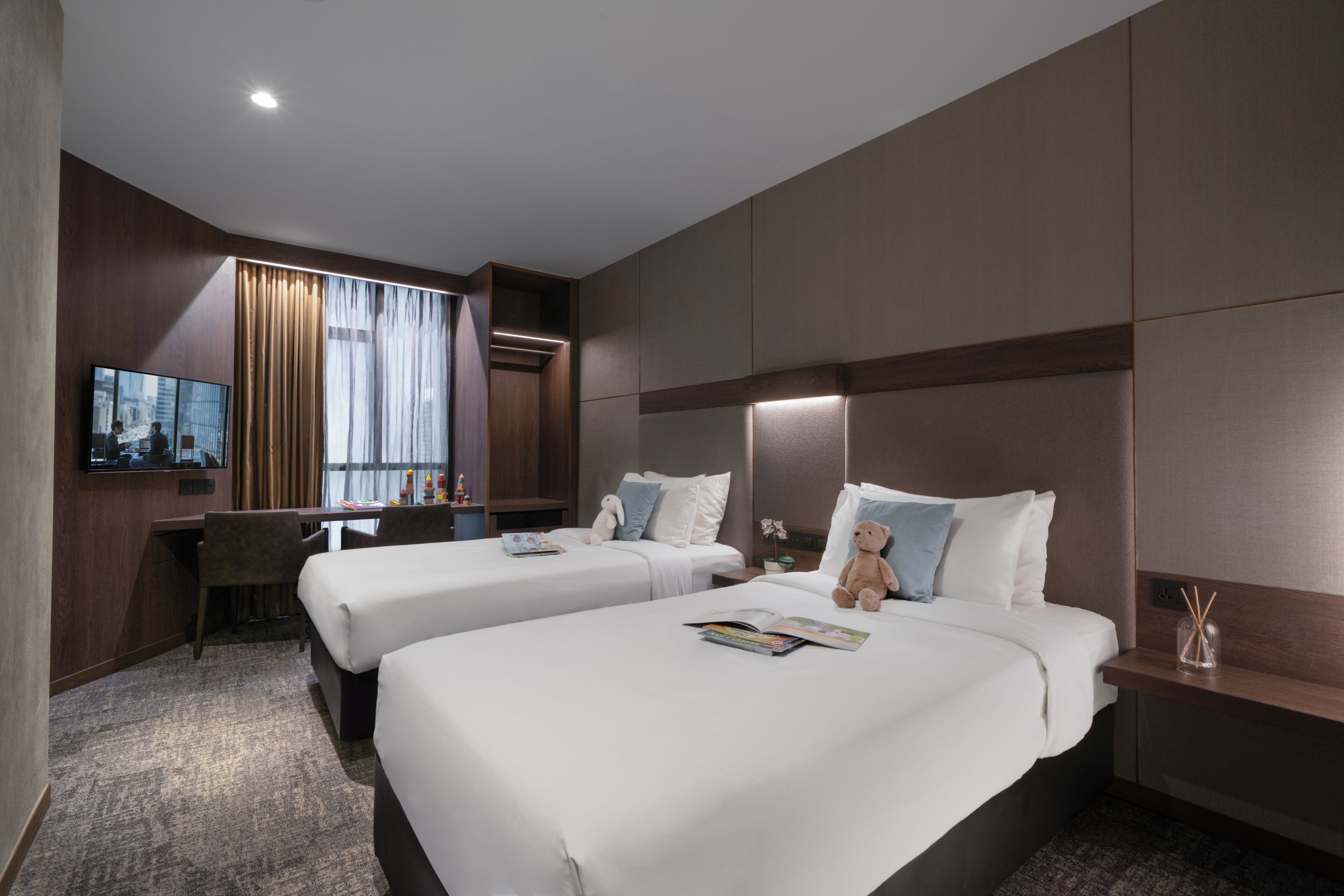 A hotel room with two beds, a desk, a TV, and a large window with curtains.