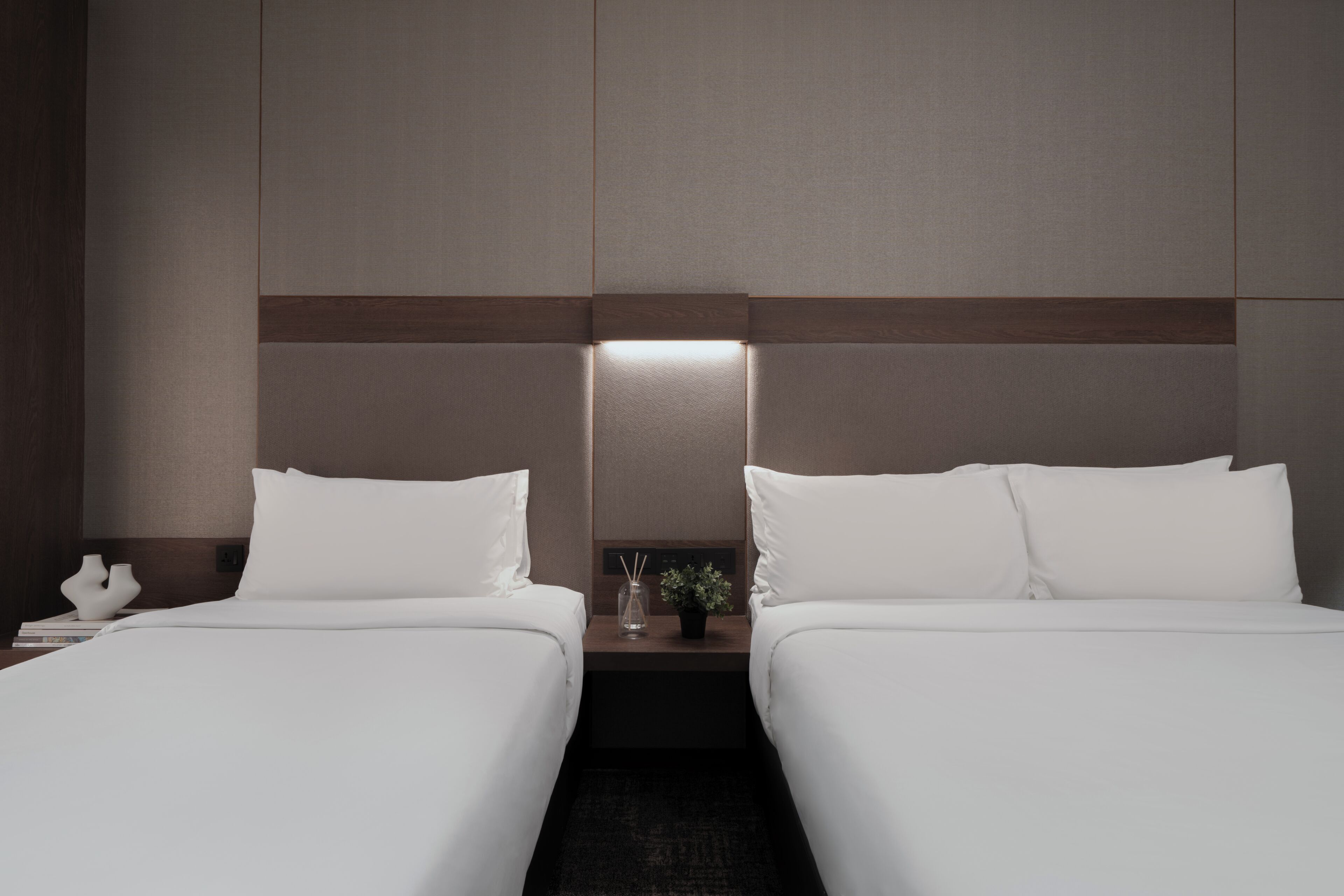 A hotel room with two single beds, a nightstand with a lamp, and a small table with a plant.