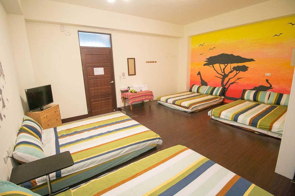 A hotel room with two beds, a television, a small table, and a mural of a sunset with giraffes.
