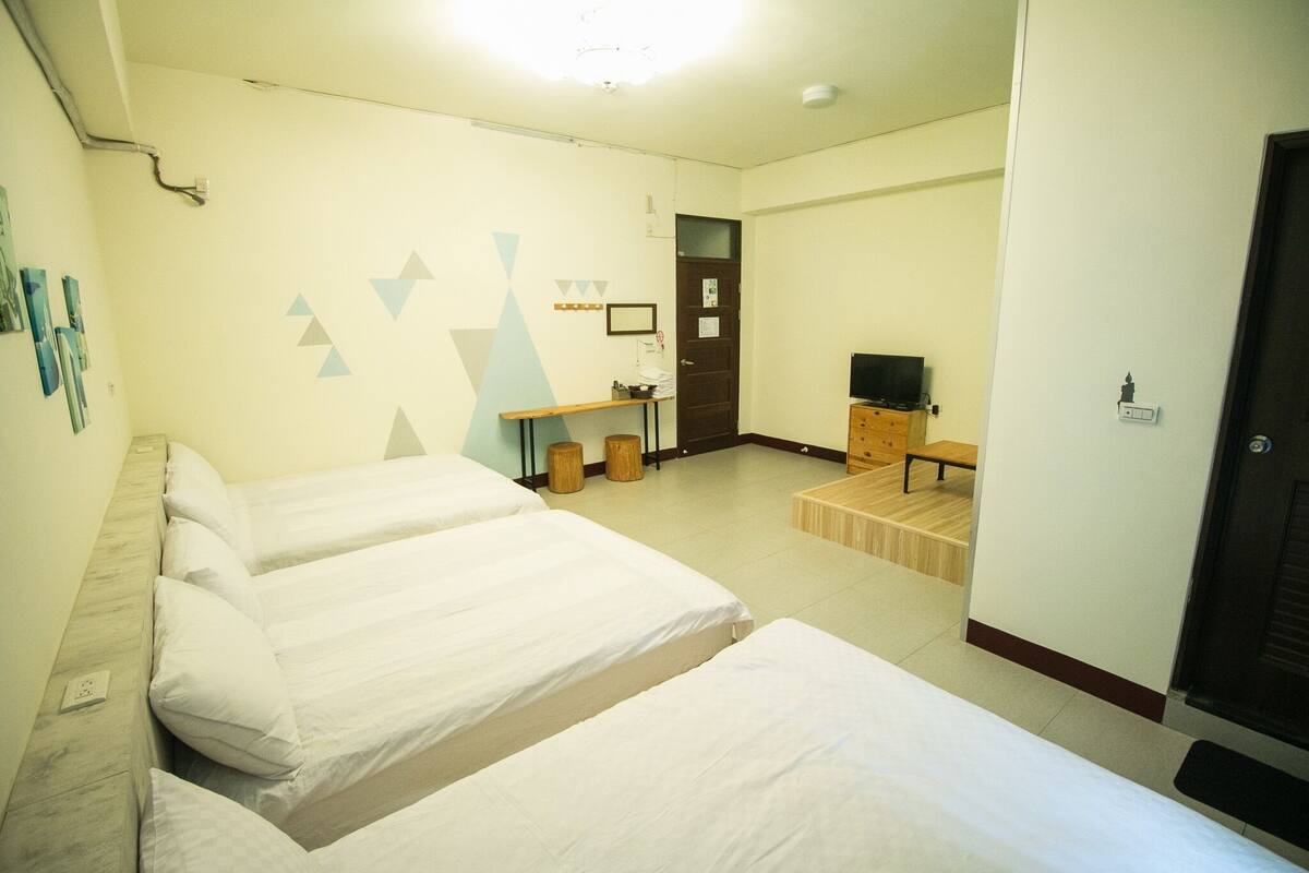 A hotel room with three beds, a wooden bench, a television, and a door.