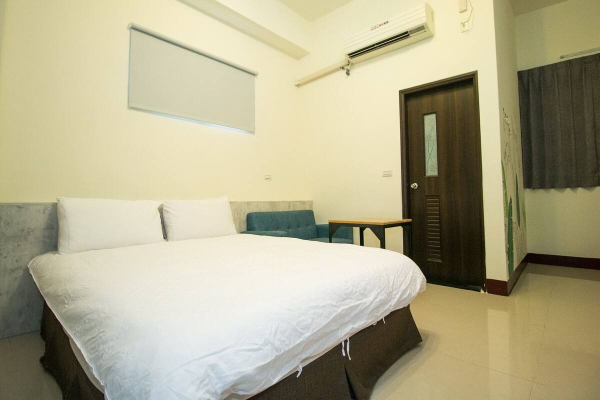 A hotel room with a large bed, a small table, a chair, and a door.