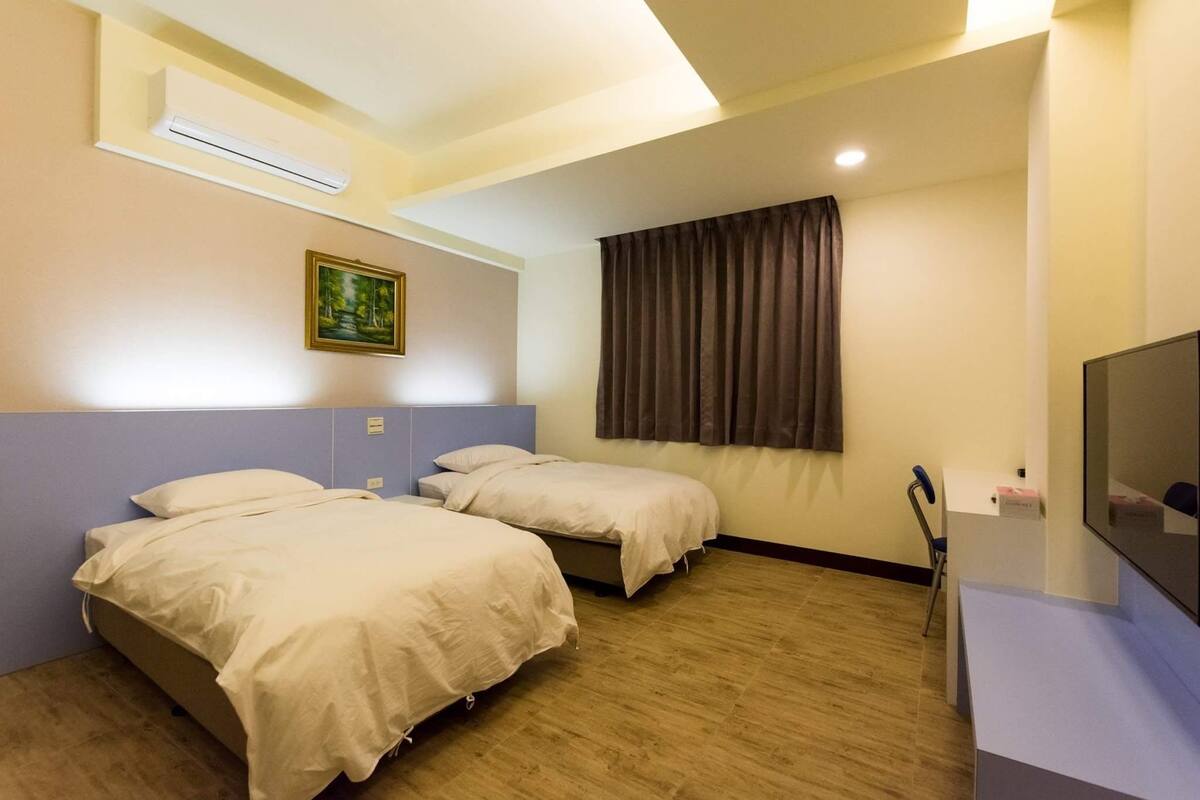 Habitación de hotel con dos camas individuales, un televisor de pantalla plana, un cuadro en la pared y una ventana con cortinas.