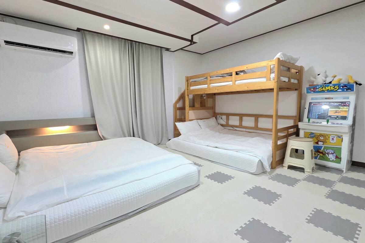 Una habitación con literas, una cama individual, un equipo de aire acondicionado, una mesita con una consola de videojuegos y un suelo con un estampado gris y blanco.