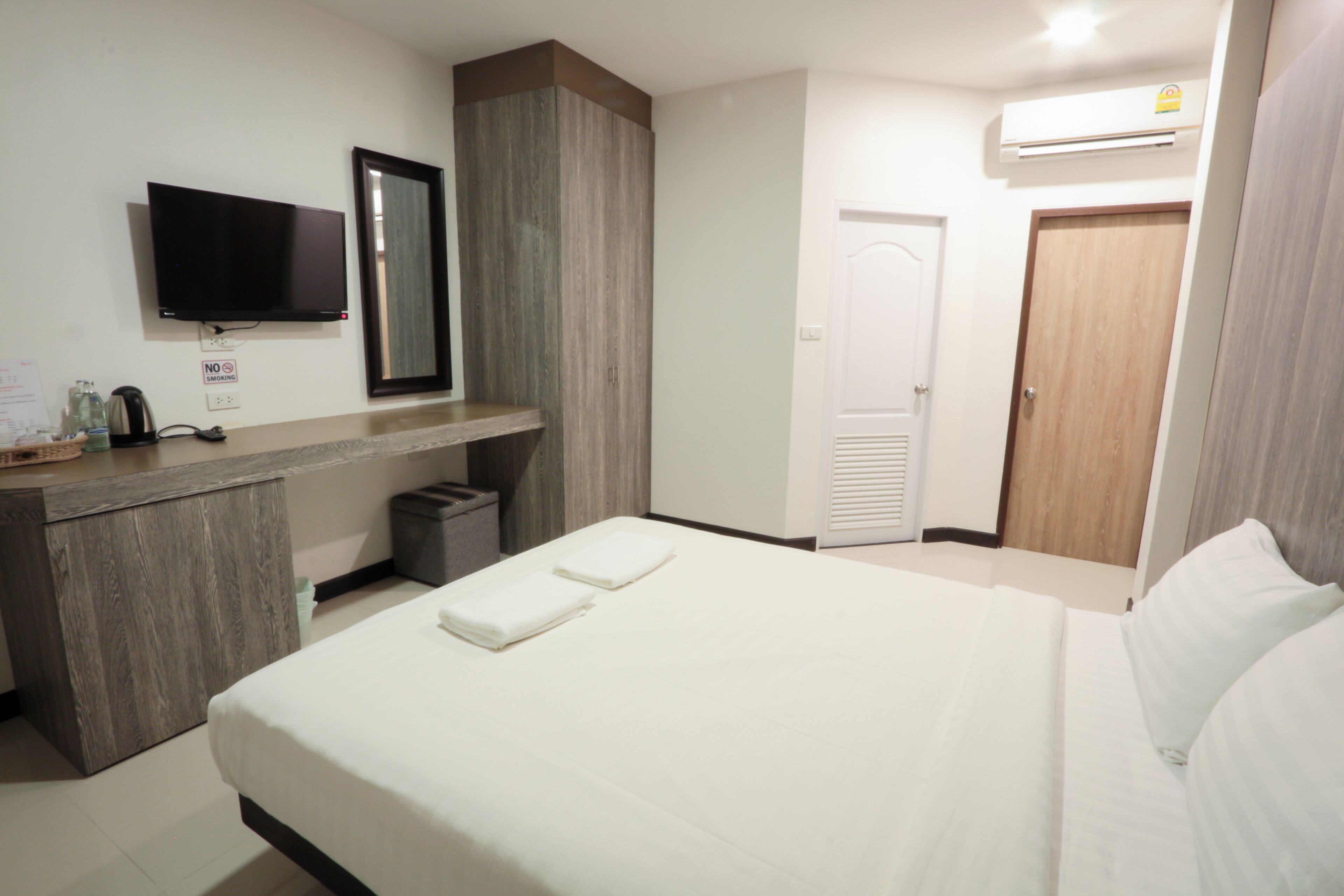 A hotel room with a bed, a TV mounted on the wall, a mirror, a wooden cabinet, a trash bin, and an air conditioning unit.