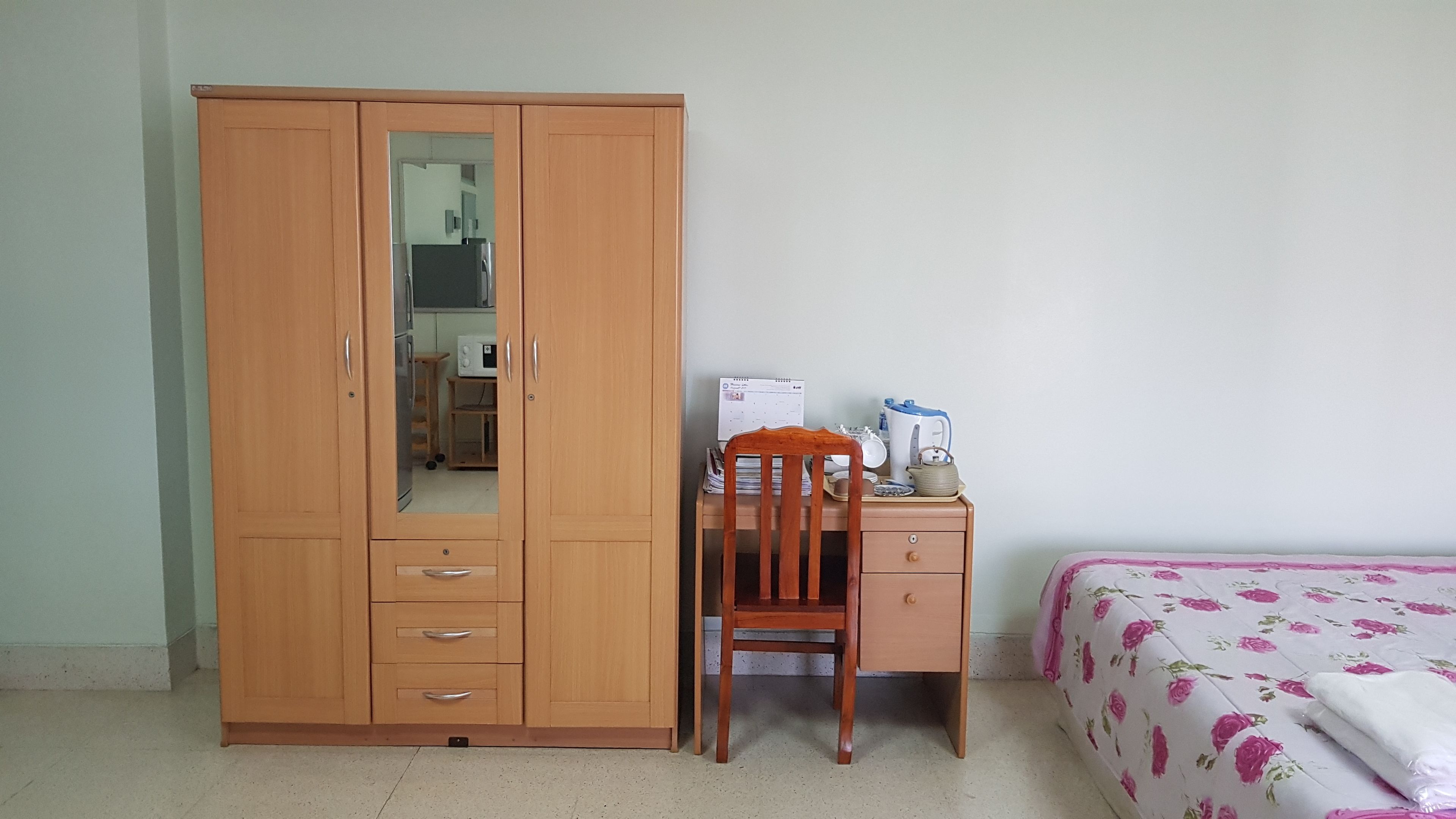 A bedroom with a wooden wardrobe, a desk with a chair, a bed with floral bedding, and a calendar on the wall.