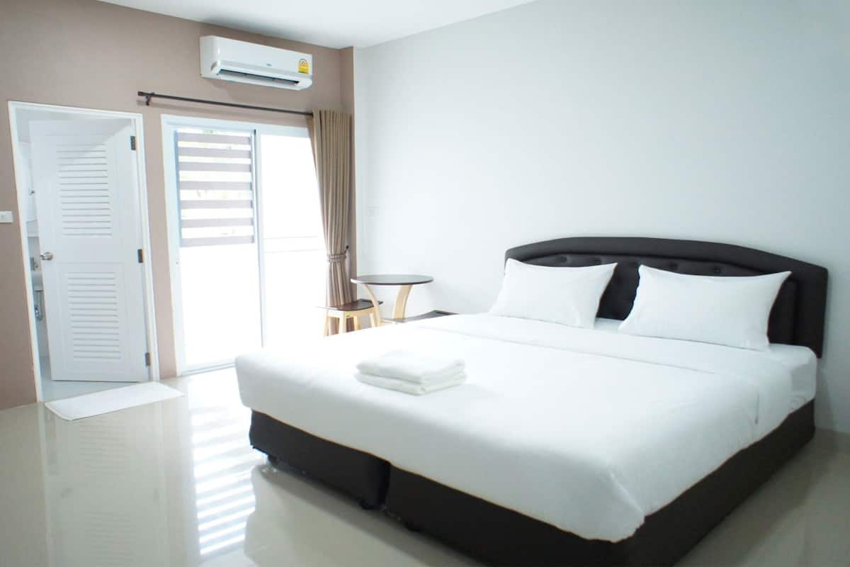 A hotel room with a bed, a bedside table, a chair, a door, and an air conditioning unit.