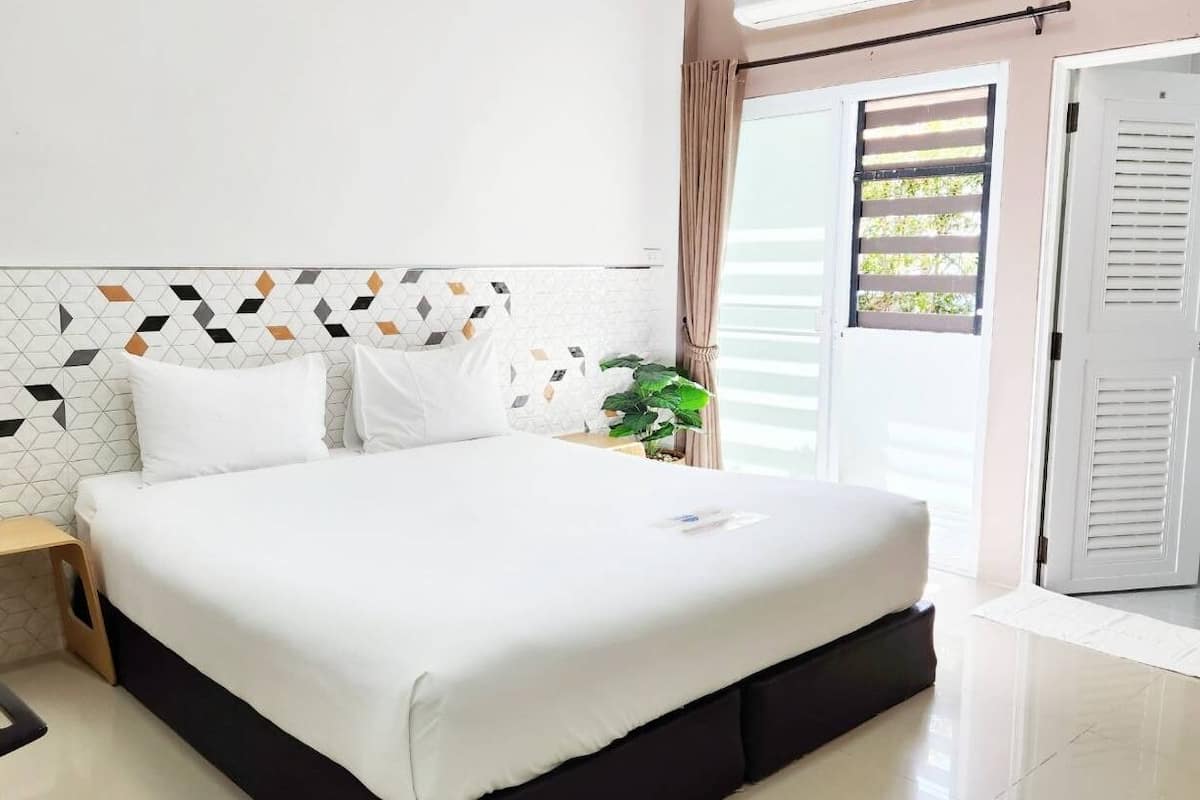 A neatly made bed with white bedding, a small wooden table with a chair, and a potted plant on the nightstand.