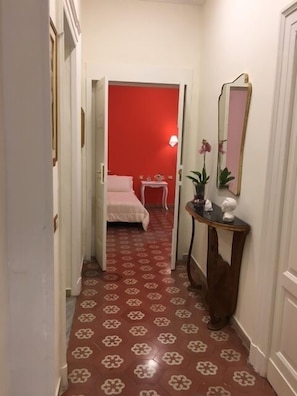 Un pasillo estrecho con piso estampado que conduce a una habitación con una pared de acento roja. Se ve una cama en la habitación, además de una mesita de noche con una lámpara y una planta en maceta.