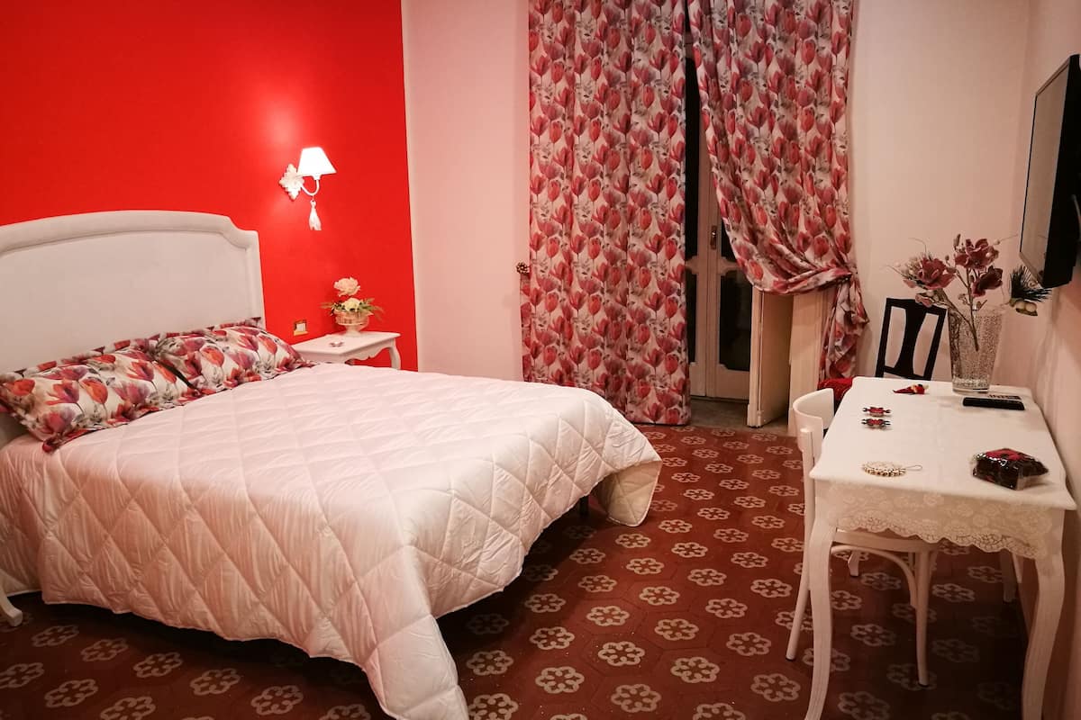 Un dormitorio con una pared roja, una cama blanca, una mesita con un jarrón y una silla.