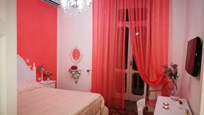 Un dormitorio con cama, mesita de noche, espejo, cómoda, escritorio con silla, candelabro y ventana con cortinas rojas.