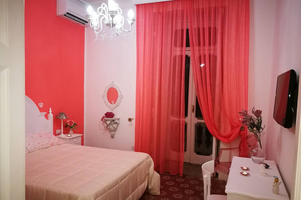 Un dormitorio con cama, mesita de noche, espejo, cómoda, escritorio con silla, candelabro y ventana con cortinas rojas.