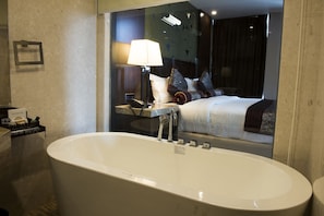 A modern bathroom with a large freestanding bathtub, a bedside table with a lamp, and a mirror reflecting a neatly made bed with patterned pillows.