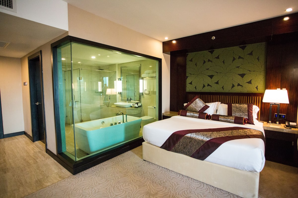 A modern hotel room with a large bed, a glass-enclosed bathroom, and a decorative wall panel.