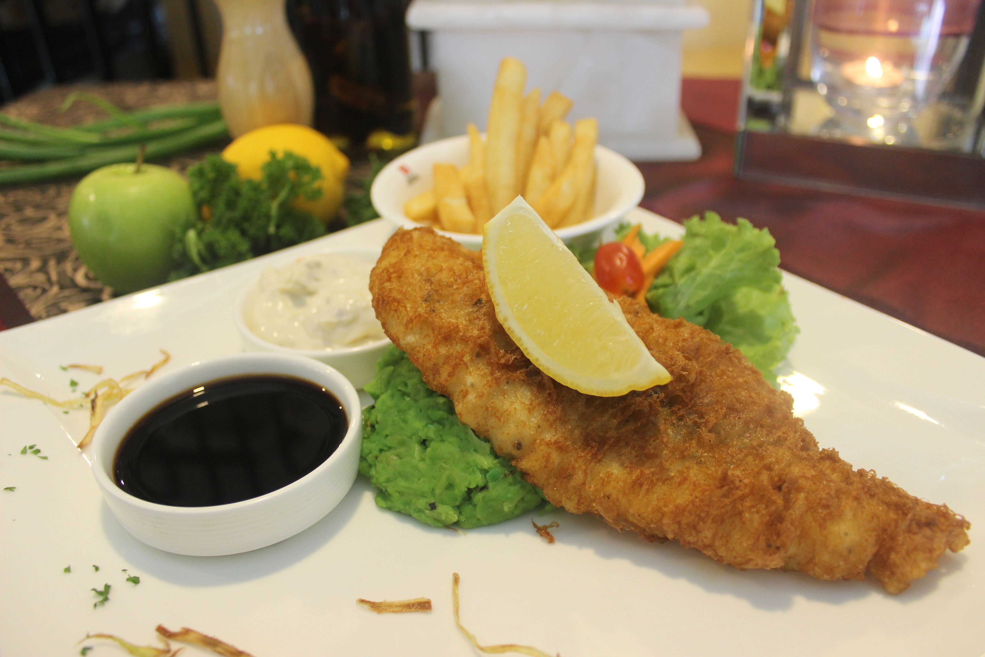 Une assiette contenant un plat pané et frit, probablement du poisson, accompagné d’une tranche de citron, d’une portion de frites et d’un petit bol de sauce.