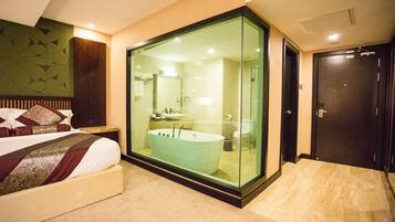 Una habitación de hotel moderna con una amplia bañera acristalada, una zona de ducha separada y una cama con ropa de cama estampada impecablemente ordenada.