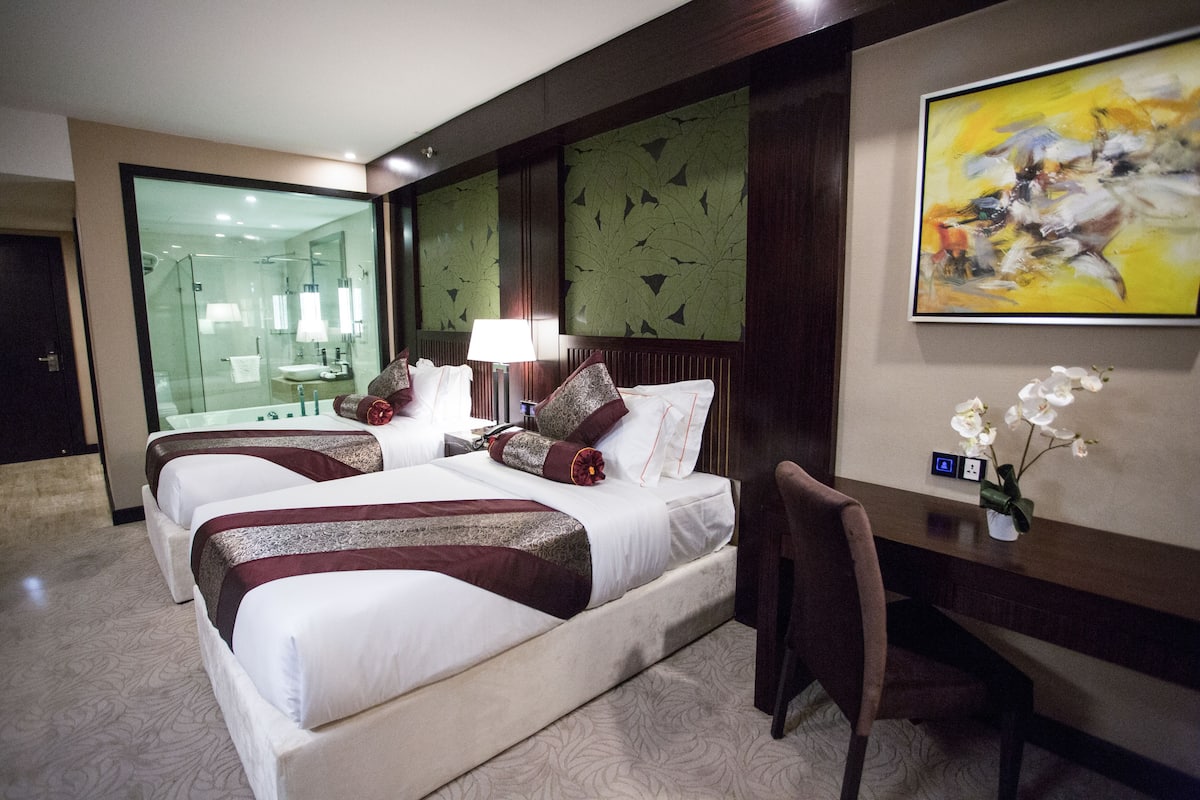 A hotel room with two beds, a desk, a chair, a desk lamp, a vase with flowers, and a painting on the wall.