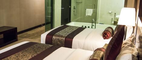 Habitación de hotel con dos camas, baño con cabina de ducha de vidrio y mesita de noche con lámpara.