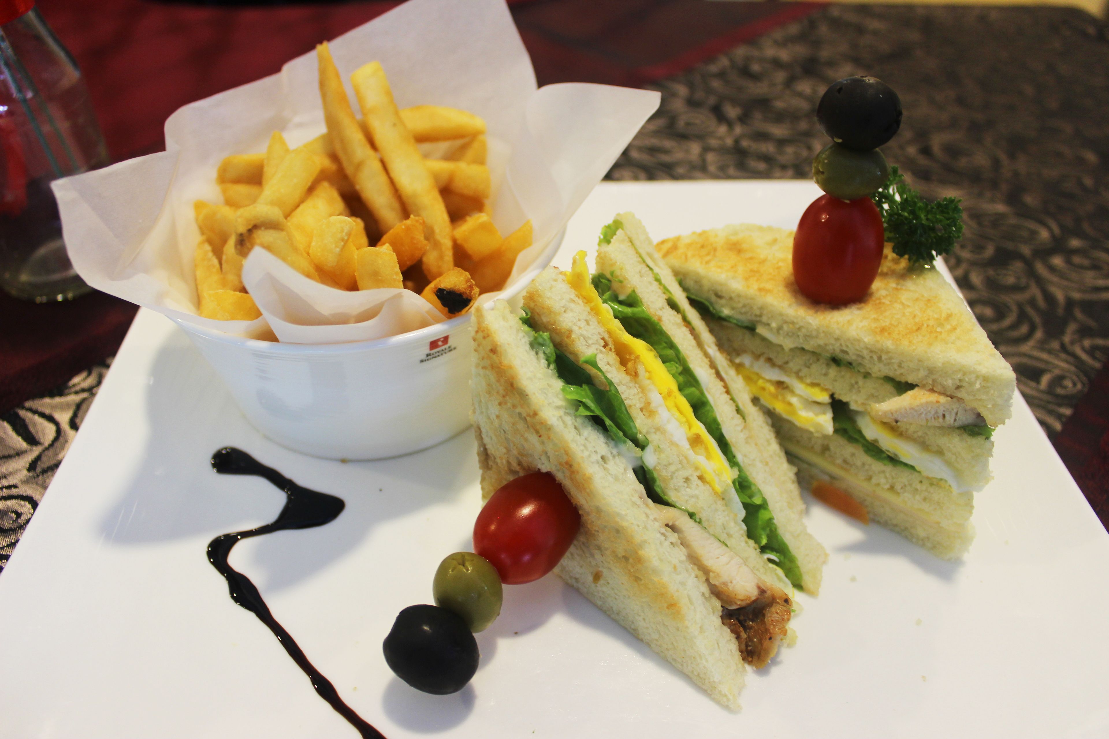 A sandwich with lettuce, tomato, and egg on a white plate, accompanied by a small bowl of fries and a few olives.