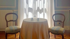 Una habitación con una mesa redonda, dos sillas, un ventanal con cortinas translúcidas y un radiador.
