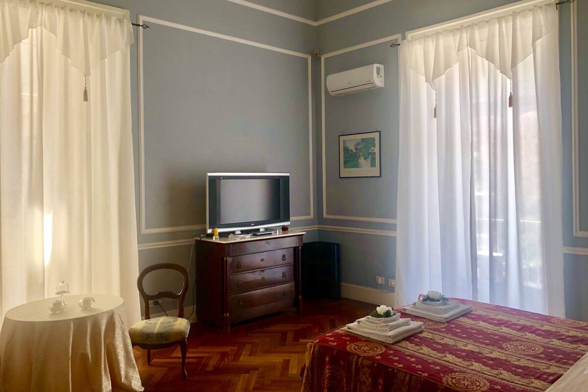 Una habitación de hotel con un televisor sobre un aparador de madera, una cama con colcha roja y dorada, y un cuadro en la pared.