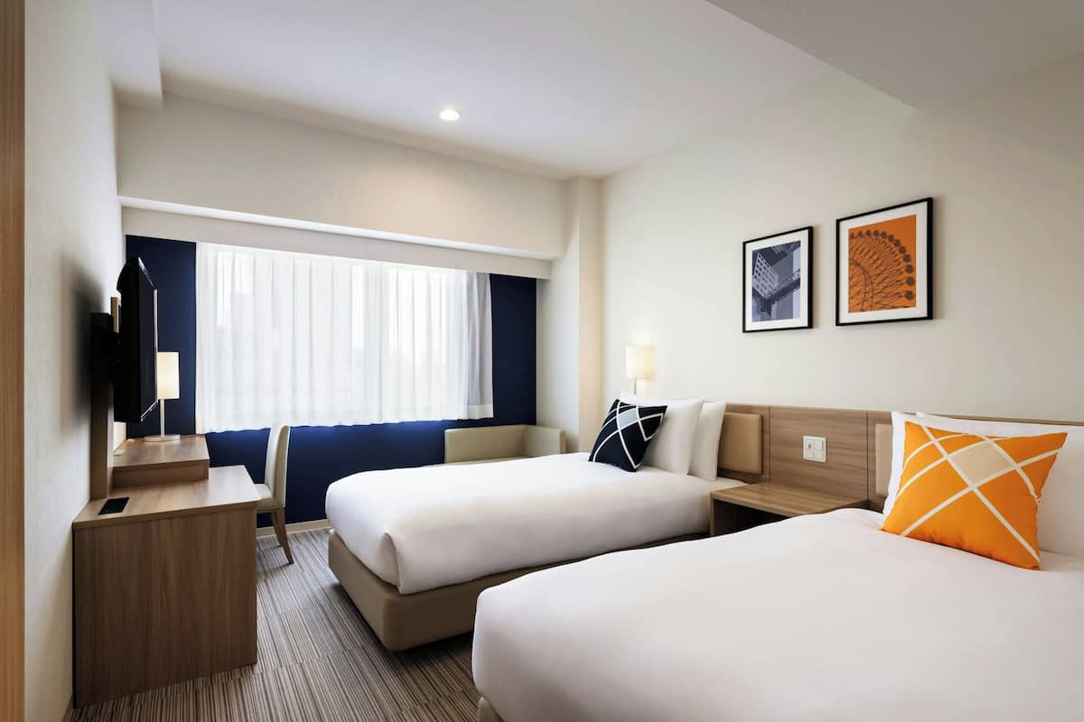 Une chambre d’hôtel avec deux lits, un bureau, une chaise et un téléviseur. Deux tableaux encadrés sont accrochés au mur.