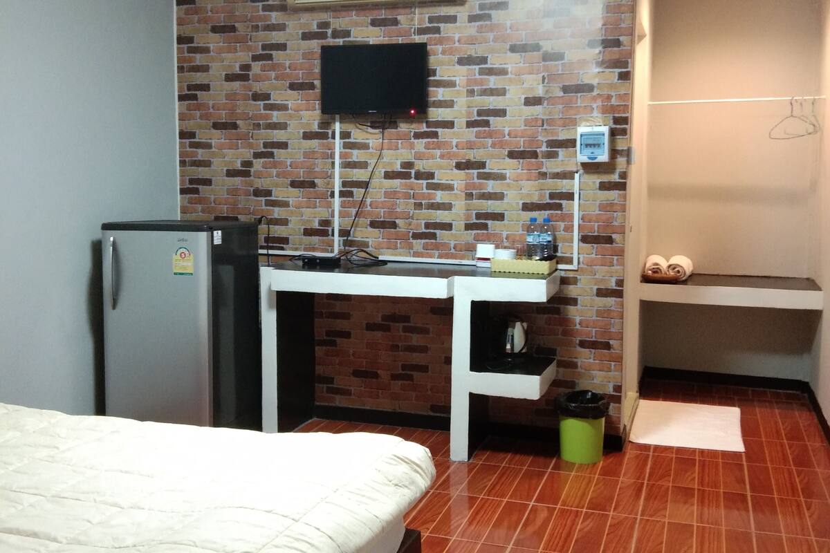 Quarto de hotel com cama, escrivaninha com televisão de tela plana, frigorífico, parede com padrão de tijolos, chão em azulejos e casa de banho pequena com lavatório e suporte para toalhas.
