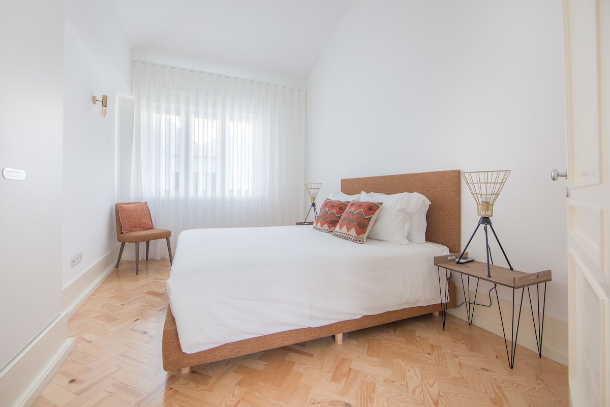 A bedroom with a wooden bed, white bedding, a chair, a bedside table with a lamp, and a window with sheer curtains.