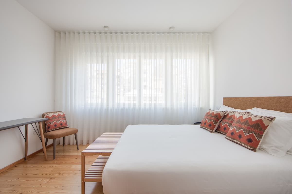 A hotel room with a bed, a desk, a chair, and a window with sheer curtains.