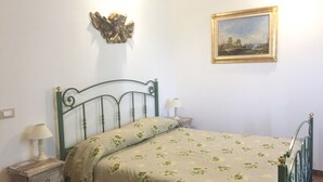 Un dormitorio con cama, mesita de noche, lámpara, ángel decorativo montado en la pared y un cuadro de paisaje enmarcado.