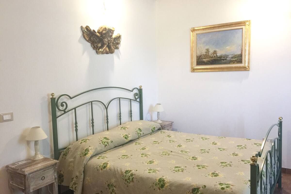 A bedroom with a bed, a nightstand, a lamp, a wall-mounted decorative angel, and a framed landscape painting.