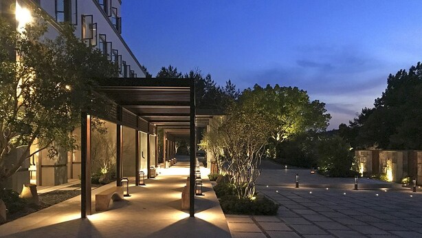 A well-lit outdoor corridor with a covered seating area, surrounded by trees and plants, leading to a building entrance. - The Lucid Resort (Taizhou)