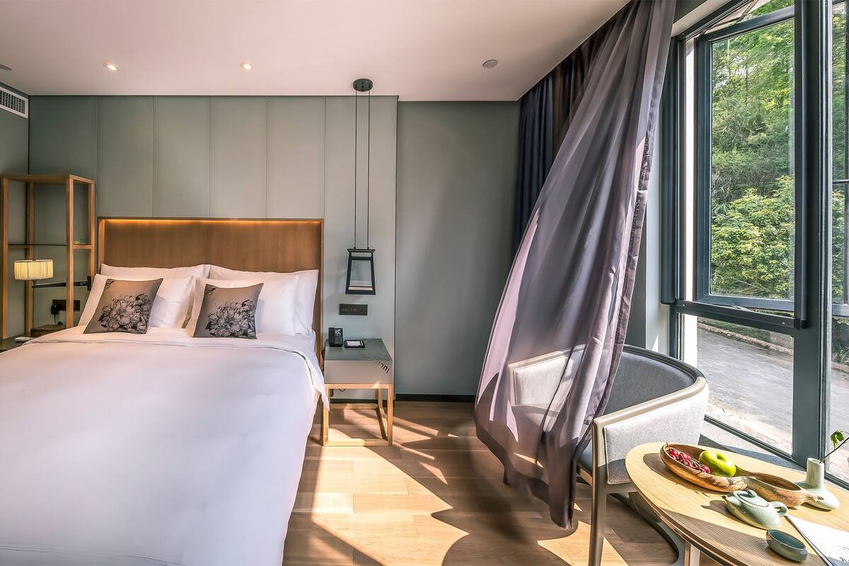 A modern hotel room with a large bed, bedside tables, a bedside lamp, a small table with fruits and a teapot, and a view of greenery through the window.