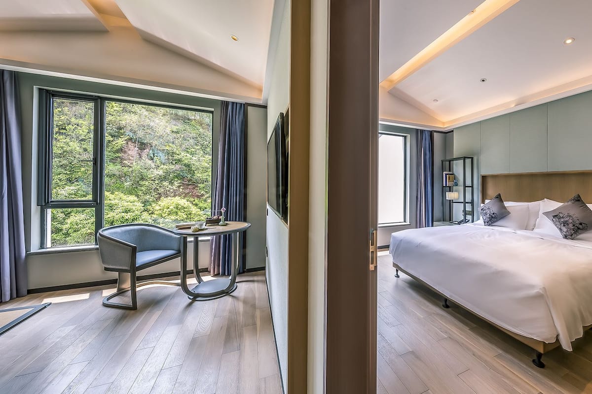 A modern hotel room with a large bed, a desk, and a chair. There is a window with a view of trees.