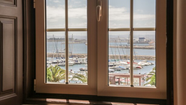 Vista desde una ventana de un puerto deportivo con barcos amarrados, cielo despejado y un paisaje urbano lejano.