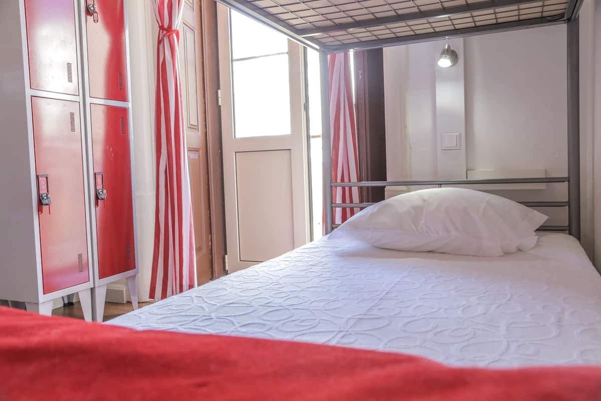 A single bed with a white pillow and a red blanket, a red and white striped curtain, and a red metal wardrobe with a lock.