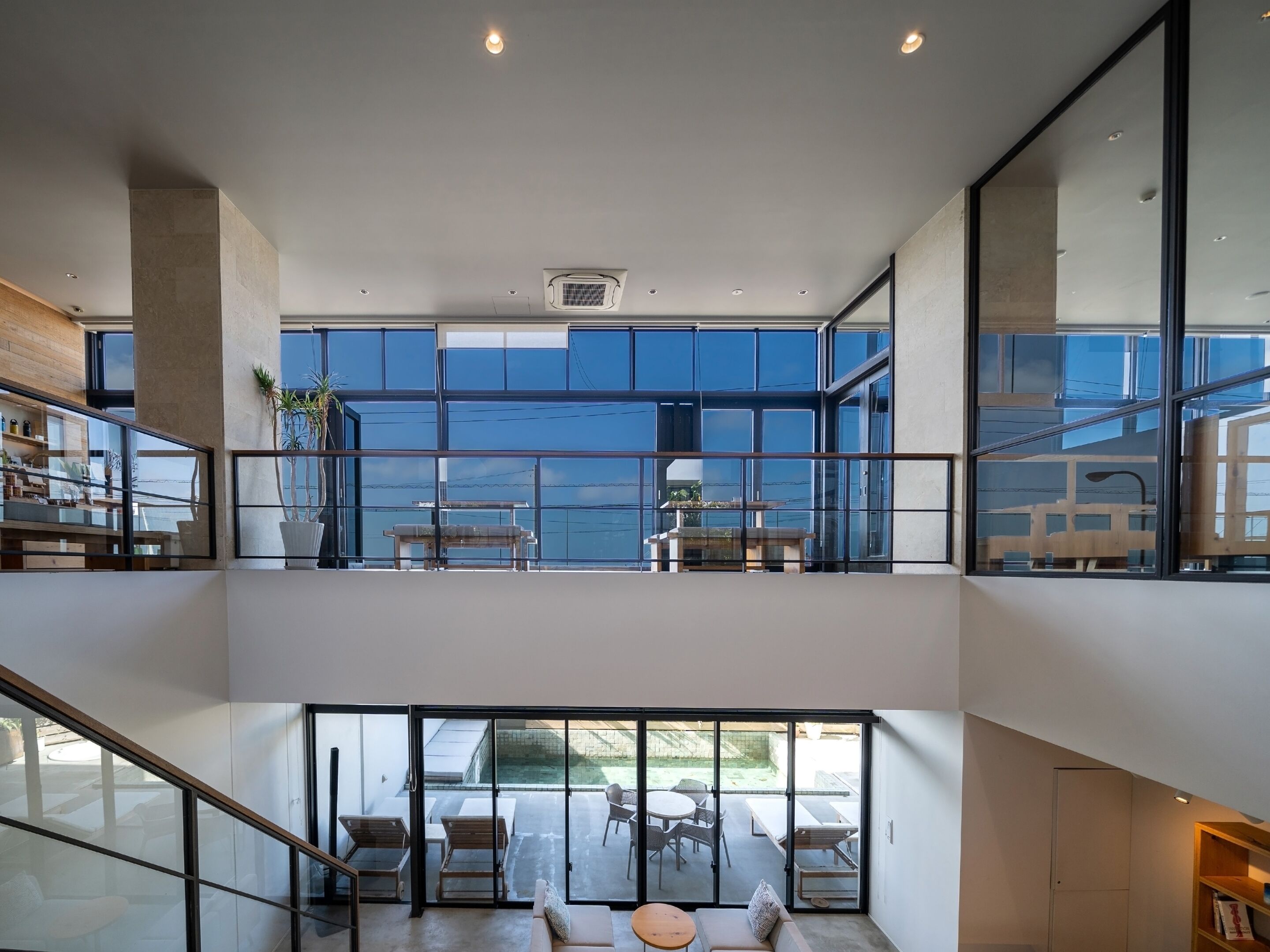A modern interior with a glass balcony overlooking a pool, a dining area with outdoor seating, and a staircase connecting the levels.