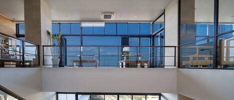 Un interior moderno con un balcón de cristal con vistas a una piscina, un área de comedor con mobiliario exterior y una escalera que conecta los niveles.